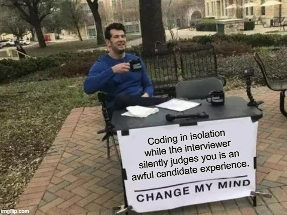 Man sitting behind a desk with a sign reading "change my mind: coding in isolation while the interviewer silently judges you is an awful candidate experience".